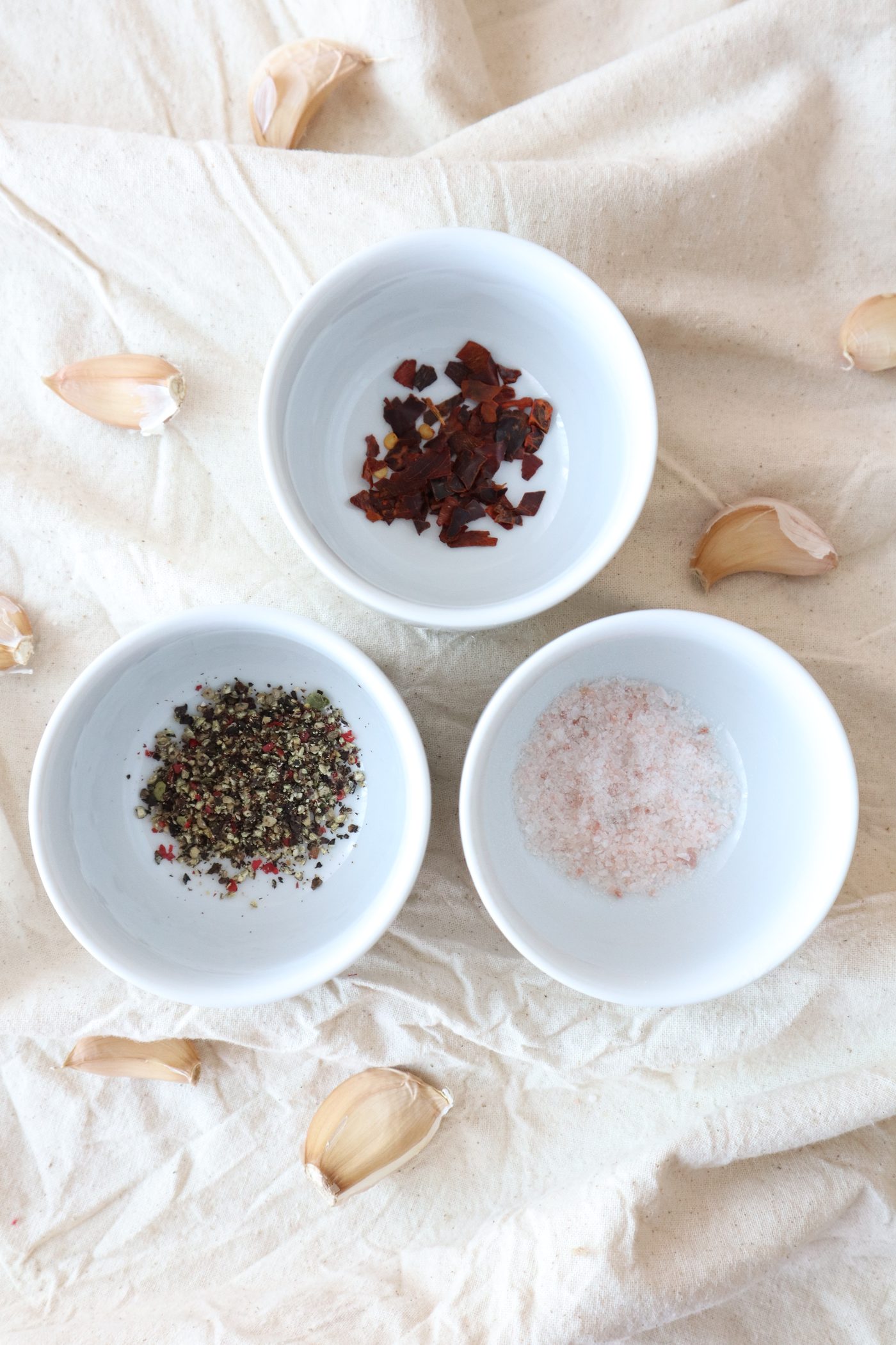 Three small bowls of chili flake, cracked pepper, and pink salt on natural linen