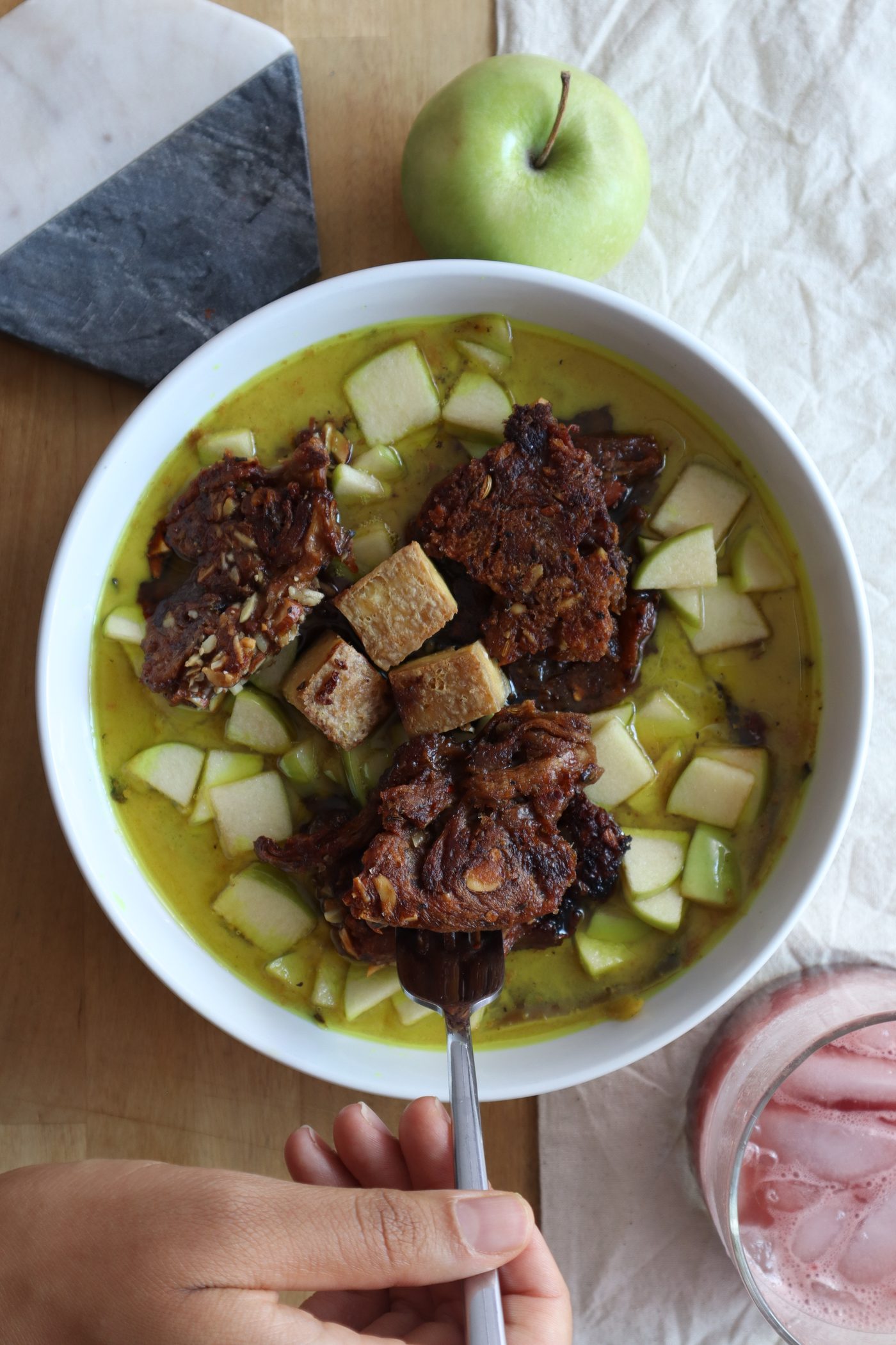 Hand lifting a bite of tofu from the golden broth bowl with apple and avocado