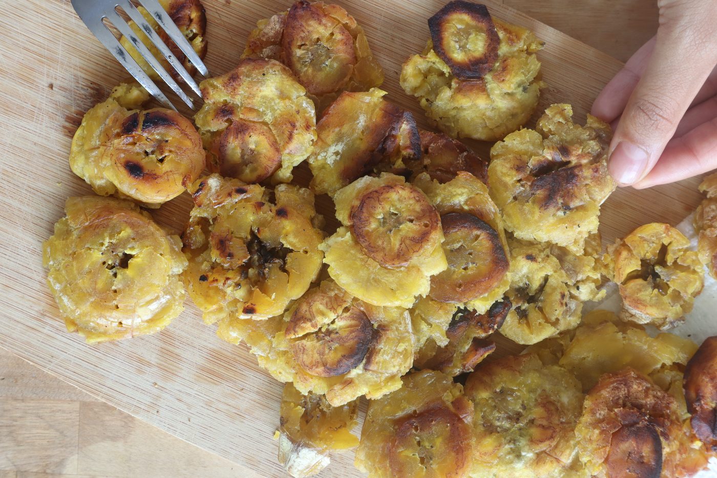 Overhead pile of golden fried tostones on a wood board, fork and hand reaching in